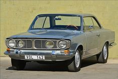 an old grey car parked in front of a yellow wall