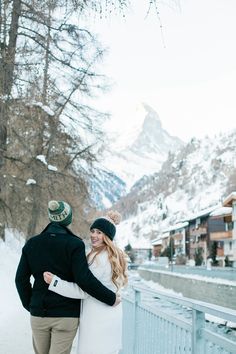 a man and woman are kissing in the snow by a fence with mountains behind them