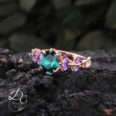 a green ring with pink and purple stones on top of a black stone surface next to some rocks