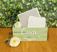 a wooden box with some cards in it on a table next to flowers and greenery