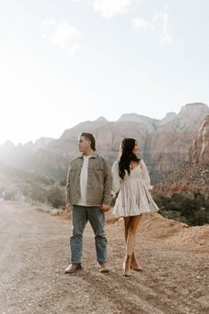 a man and woman holding hands while standing on the side of a road in front of mountains