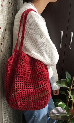 a woman is holding a red crocheted bag in front of a wall with plants