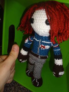 a crocheted doll with red hair is in a green box and someone is holding it up to the camera