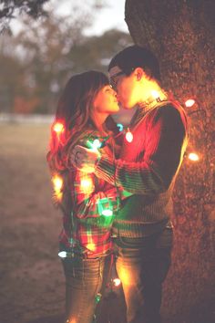 two people standing next to each other near a tree with christmas lights on the branches