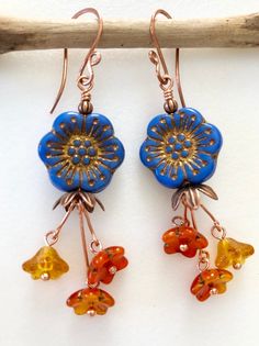 "Colorful Czech glass flower beads in cornflower blue with orange and topaz flower dangles on handmade copper ear wires. Cornflower blue Czech glass wild rose is 17mm with gold/copper Picasso etching accented with copper bead caps. Darling little mandarin orange/gold cup flowers and topaz bellflowers dangle on handmade bright copper stems. Rustic flower dangles hang on handmade hammered German copper ear wires. Total drop is 2 1/2\"." Blue Czech Glass Flower Earrings, Blue Bohemian Flower Earrings, Czech Glass Bead Earrings, Picasso Flowers, Czech Beads Jewelry, Blue Flower Earrings, Czech Glass Jewelry, Glass Bead Earrings, Earrings Inspiration