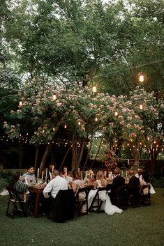 a group of people sitting around a dinner table under a tree with lights on it