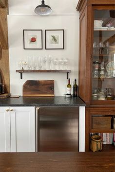 a kitchen counter with wine glasses and bottles on the shelf above it, next to a wooden cabinet