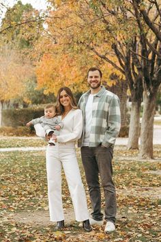 a man and woman holding a baby in their arms while standing next to trees with leaves on the ground