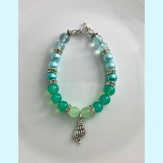 Experience the enchanting beauty of our Ombre Green Teal Bracelet adorned with a delicate Silver Shell Charm and a silver toggle clasp. Handmade with love, this bracelet exudes a boho chic vibe and captures the essence of beachy vibes. The ombre design, with varying shades of green and teal beads, creates a mesmerizing and captivating accessory. Each bead is carefully selected to create a seamless transition of colors, adding depth and charm to the bracelet. The silver shell charm adds a touch of coastal elegance and whimsy, evoking memories of sunny beach days and tranquil ocean waves. It serves as a reminder of the serenity and beauty of the seaside. Crafted with meticulous attention to detail, this bracelet is not only a stylish accessory but also a reflection of your unique style. The Bohemian Green Crystal Bracelet For Beach, Adjustable Ocean-inspired Beaded Bracelets With Lobster Clasp, Adjustable Ocean-inspired Charm Bracelet With Lobster Clasp, Green Crystal Bracelet With Round Beads For Beach, Handmade Dangle Bracelets For Beach, Adjustable Bracelet With Lobster Clasp For Vacation, Handmade Turquoise Crystal Bracelet For Beach, Ocean-inspired Adjustable Charm Bracelet As Gift, Ocean-inspired Adjustable Charm Bracelet Gift