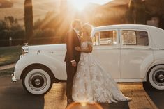 a bride and groom standing in front of an old white wedding car with the sun behind them