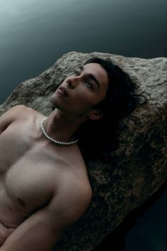 a shirtless man laying on top of a rock next to the water and wearing a pearl necklace