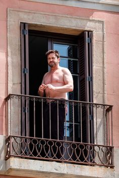 a shirtless man standing on a balcony looking out the window with his hands in his pockets