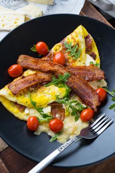 an omelet with bacon, tomatoes and greens on a black plate next to a fork