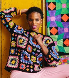 a woman posing in front of a colorful wall with her hands on her head, wearing pink pants and a crocheted jacket