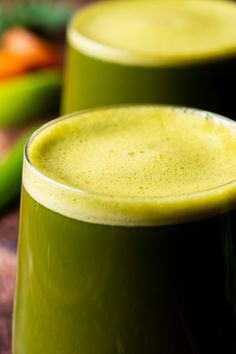 two glasses filled with green smoothie sitting on top of a wooden table next to vegetables