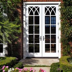 a white double door in front of a brick building with flowers and bushes around it