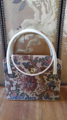 "Lovely 60's tapestry hand bag. Tapestry design fabric on both sides and closing flap. Metal closure at front. White leather (not sure if real or faux) on handles, trim and on the bottom. The bottom has little metal feet. Inside is silky mink coloured fabric with two sides and one zipped pocket in the middle. This bag is in pretty much new condition!! 9 1/2\" down, 11 1/2\" across, 5\" width, 21\" long handles each." Vintage Tapestry Shoulder Bag With Leather Handles, Vintage White Satchel With Handles, Retro Rectangular Tapestry Bags, Vintage Tapestry Satchel With Top Handle, Vintage Rectangular Fabric Bag, Vintage Tapestry Satchel With Top Carry Handle, Vintage Rectangular Fabric Shoulder Bag, White Handles, Tapestry Handbags