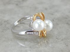 This cocktail ring makes quite a statement! The unadorned, highly polished gold compliments the bright white cultured pearls. Channel set diamonds provide a little bit of glitter, but the smooth setting, with no prongs, retains the modernist, contemporary feel of the piece. Perfect for a night out on the town, comfortable for every day. Metal: 14K Yellow and White Gold Gem: (2) Bright White Pearls, Cultured Gem Measurements: 7mm Round Accents: 8 Diamonds totaling .08 Carat Size of Ring: 5.25 SKU Classic White Pearl Ring With Diamond Accents, White Pearl Rings With Diamond Accents, White Diamond Ring With Diamond Accents, Classic Pearl Ring With Diamond Accents For Anniversary, Diamond Pearl Ring With Polished Finish For Anniversary, White Gold Akoya Pearl Diamond Anniversary Ring, White Gold Akoya Pearl Anniversary Ring, White Akoya Pearl Rings With Diamond Accents, White Pearl Ring With Diamond Accents For Anniversary