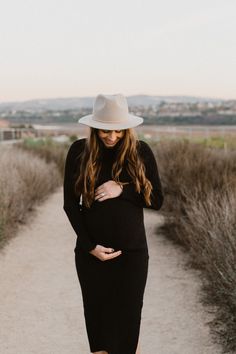 a pregnant woman in a black dress and hat walks down a path with her hands on her stomach