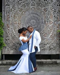 a man and woman dressed in blue and white dance together on the sidewalk near a carved wooden wall