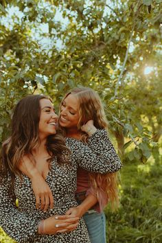 two women hugging each other while standing in front of some trees and bushes with the sun shining on them