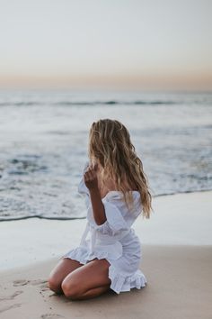 a woman sitting on top of a sandy beach next to the ocean with her hand in her mouth