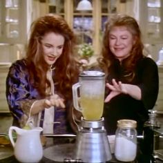 two women standing in front of a blender on a kitchen counter, one is pouring something into the other