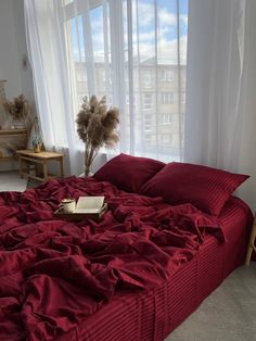 an unmade bed with red sheets and pillows in front of a large window overlooking the city