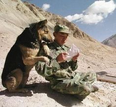 a man kneeling down next to a dog on top of a dirt field with mountains in the background