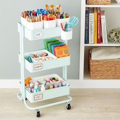 a cart filled with lots of crafting supplies on top of a wooden floor next to a bookshelf