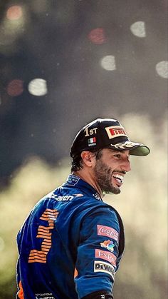 a man wearing a hat and smiling while standing on top of a dirt field with trees in the background