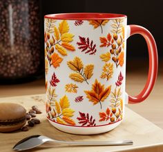 a coffee mug with autumn leaves painted on it next to a spoon and some coffee beans