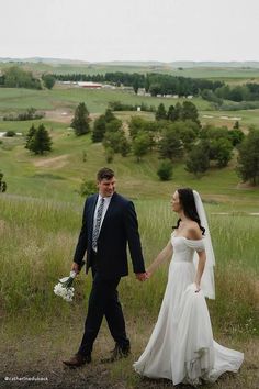 a bride and groom holding hands while walking through the grass in front of an open field