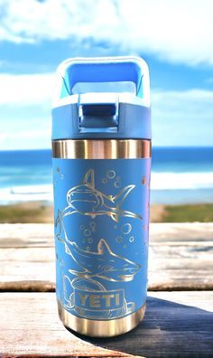 a blue and gold insulated water bottle sitting on top of a wooden table next to the ocean