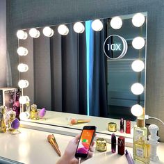 a person holding a cell phone in front of a vanity mirror with lights on it