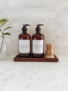 two bottles of hand soap sitting on top of a wooden tray next to a brush