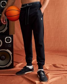 A person dressed in black Psycho Bunny MENS LEVY ESSENTIAL FLEECE SWEATPANT - B6P721D200 and black sneakers holds an orange basketball. They stand indoors against an orange curtain backdrop beside a set of large speakers. The fleece sweatpants feature a small bunny logo, and the waistband reads Psycho Bunny. Bunny Man, Oversized Tee Shirt, Tennis Shop, Swim Pants, Polo Tees, Swimwear Bottoms, Sports Tees, Top Graphic Tees, Long Sleeve Sweatshirts