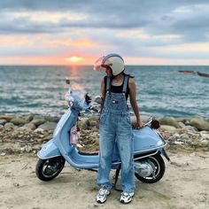 a woman in overalls standing next to a scooter on the beach at sunset