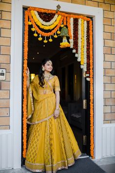 a woman standing in front of a doorway wearing a yellow lehenga and orange garland