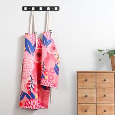 two pink and blue flowered bags hanging from hooks on a white wall next to a dresser