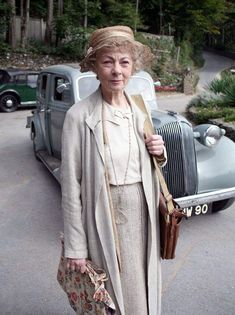 an older woman is standing in front of a vintage car and holding her handbag