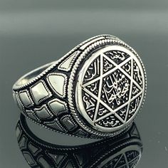 a silver ring with an intricate design on the front and side, sitting on a reflective surface