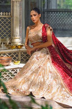 A gold tissue lehnga and choli features detailed zardoze and resham embroidery and is paired with a contrasting crimson cotton net dupatta adorned with gold zardoze borders and sequins. Gold Sharara In Raw Silk With Intricate Embroidery, Gold Intricate Embroidered Raw Silk Sharara, Gold Dola Silk Sets With Mirror Work, Gold Raw Silk Sharara With Intricate Embroidery, Unstitched Lehenga With Intricate Embroidery For Festive Occasions, Festive Lehenga With Intricate Embroidery Unstitched, Gold Lehenga With Dupatta In Raw Silk, Gold Raw Silk Lehenga With Dupatta, Gold Saree Dress With Mirror Work