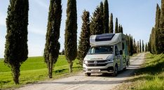 an rv parked on the side of a dirt road next to tall trees and grass