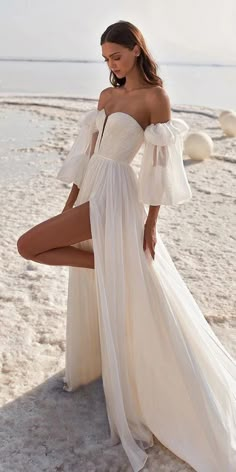 a woman posing on the beach wearing a white dress and high heeled sandals with her legs crossed