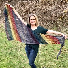 a woman holding up a multicolored crocheted shawl in the grass