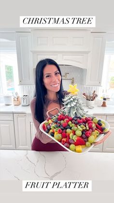 a woman holding a platter full of fruit in front of a christmas tree on the counter