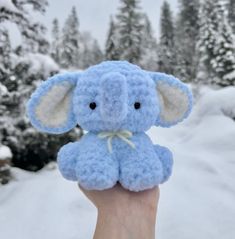 a small blue stuffed elephant sitting on top of a persons hand in front of snow covered trees