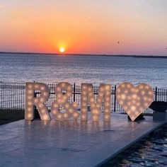 the letters r and m are lit up in front of an ocean at sunset or dawn