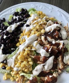 a white plate topped with a salad covered in ranch dressing and black olives next to corn on the cob